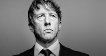 Jonathan Pie looks stern as he stands dressed in a suit.