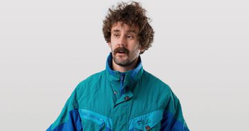 Josh Pugh, a young white man with curly brown hair, a thick moustache, and stubbly beard in a blue windbreaker jacket on a white background looking confused