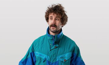 Josh Pugh, a young white man with curly brown hair, a thick moustache, and stubbly beard in a blue windbreaker jacket on a white background looking confused
