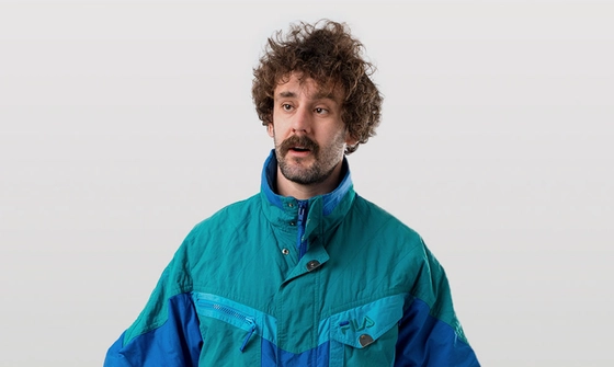 Josh Pugh, a young white man with curly brown hair, a thick moustache, and stubbly beard in a blue windbreaker jacket on a white background looking confused
