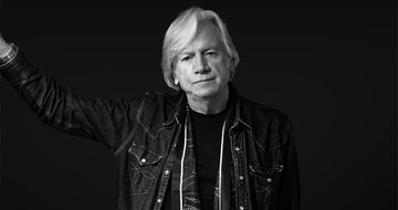 A black and white photo of Justin Hayward leaning against a wall out of shot in a denim jacket