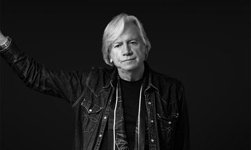 A black and white photo of Justin Hayward leaning against a wall out of shot in a denim jacket