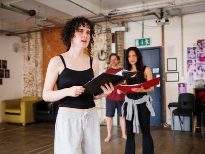 Kathleen McAuley Irvine, Jim Fish and Amy Tara rehearse a scene, standing spaced apart from each other while holding scripts.