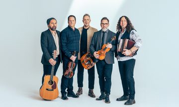 All five men stand in a line together, they all are dressed smart casual and hold their respective instruments as they stand against a white backdrop