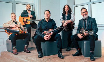Five older white men sat holding instruments in front of a white wall and windows. They're holding a hurdy gurdy, guitar, violin, accordion, and a mandolin