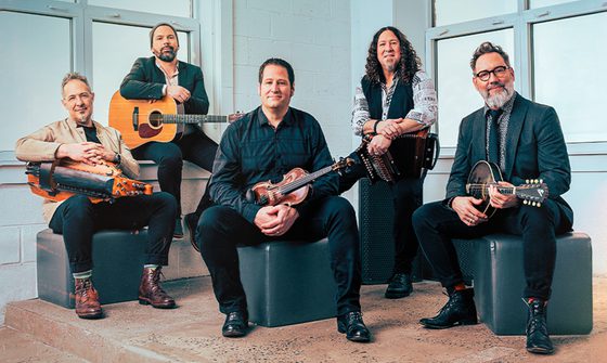 Five older white men sat holding instruments in front of a white wall and windows. They're holding a hurdy gurdy, guitar, violin, accordion, and a mandolin