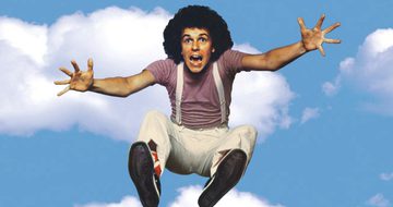 A man with a brown afro, jumps into the sky. Around him is the blue sky and fluffy white clouds. He has his arms out and is screaming, he's wearing a mauve t-shirt and white trousers with white suspenders.