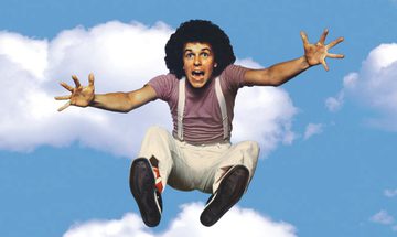 A man with a brown afro, jumps into the sky. Around him is the blue sky and fluffy white clouds. He has his arms out and is screaming, he's wearing a mauve t-shirt and white trousers with white suspenders.