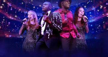 Two black men, one in an embellished black blazer and the other in a red shirt stand together holding microphones. Behind them two white women, one blonde one brunette both sing into their microphones