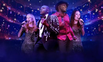 Two black men, one in an embellished black blazer and the other in a red shirt stand together holding microphones. Behind them two white women, one blonde one brunette both sing into their microphones