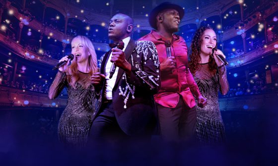 Two black men, one in an embellished black blazer and the other in a red shirt stand together holding microphones. Behind them two white women, one blonde one brunette both sing into their microphones