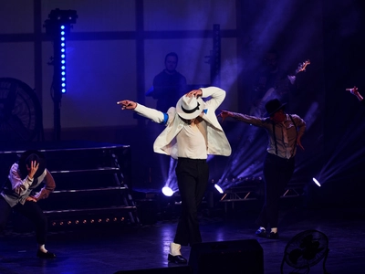 A Michael Jackson impersonator on stage dancing in a white jacket and matching hat with a group of dancers. He is pointing to the left and looking down holding his hat at the back