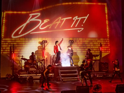 A Michael Jackson impersonator stood on stage singing in a red leather with black accents jacket and white t-shirt with a group of four dancers a small band behind with a large red neon sign reading 'Beat It!'