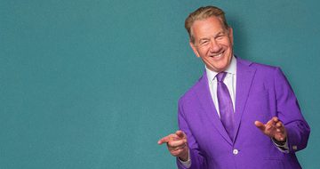 Michael Portillo wearing a purple suit against a blue-green background. He is pointing and smiling.