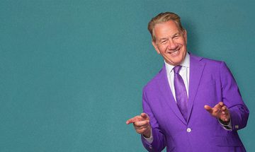 Michael Portillo wearing a purple suit against a blue-green background. He is pointing and smiling.