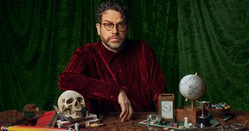 Michael Spicer in a red velvet shirt and round glasses looking very serious. In front of him is a table with lots of items including a skull that he's leaning his arm on, books, crystal ball, carriage clock, globe, board game, trophies, big pencils, and big keys
