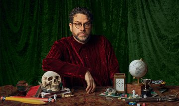 Michael Spicer in a red velvet shirt and round glasses looking very serious. In front of him is a table with lots of items including a skull that he's leaning his arm on, books, crystal ball, carriage clock, globe, board game, trophies, big pencils, and big keys
