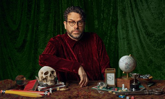 Michael Spicer in a red velvet shirt and round glasses looking very serious. In front of him is a table with lots of items including a skull that he's leaning his arm on, books, crystal ball, carriage clock, globe, board game, trophies, big pencils, and big keys