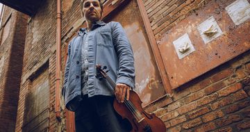 Stood in front of a brick wall outside a building Mikey Kenney holds his fiddle. He's wearing a denim jacket and jeans