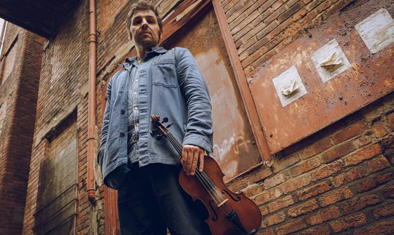 Stood in front of a brick wall outside a building Mikey Kenney holds his fiddle. He's wearing a denim jacket and jeans