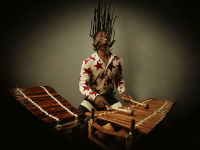 N'famady Kouyaté a young black man with long dreaded hair in a white shirt with stars playing the balafon. He is throwing his head back with his hair appearing to be stood up right