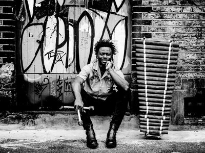 Black and white photo of N’famady Kouyaté, a young black man sat on a step with his chin resting on one hand and drum sticks in the other next to his balafon. The wall behind his is covered in graffiti