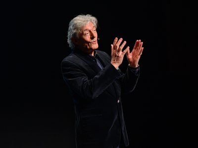 Nigel Havers speaks by a dark background, gesturing with both hands while wearing a headset microphone.