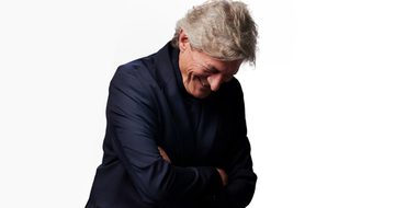 A photo of Nigel Havers in a dark blue shirt leaning forward with his arms crossed laughing
