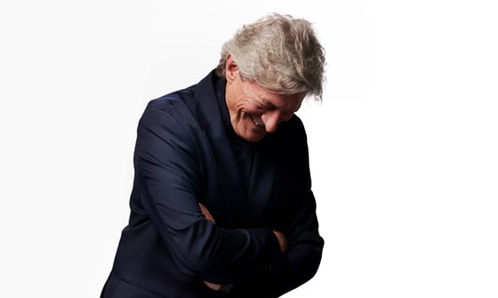 A photo of Nigel Havers in a dark blue shirt leaning forward with his arms crossed laughing