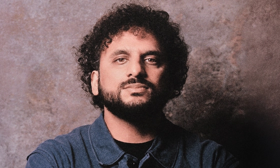 Nish Kumar looks serious as he stands against a limewashed wall. He wears a dark blue denim shirt over a black t-shirt and crosses his arms.
