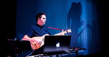 Chris Green, a white man with short brown hair in a blue shirt, is playing am acoustic guitar in front of a keyboard and computer with the ghoulish figure of Nosferatu projected on the wall behind him