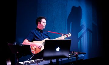 Chris Green, a white man with short brown hair in a blue shirt, is playing am acoustic guitar in front of a keyboard and computer with the ghoulish figure of Nosferatu projected on the wall behind him