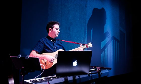 Chris Green, a white man with short brown hair in a blue shirt, is playing am acoustic guitar in front of a keyboard and computer with the ghoulish figure of Nosferatu projected on the wall behind him