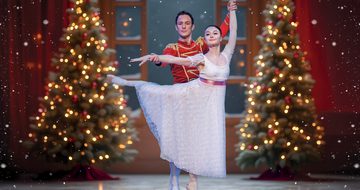 A ballerina in a white dress with pink waist ribbon in an arabesque with a man in a red toy soldier outfit holding her wrists in front of a large snowy window and Christmas trees