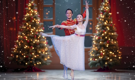 A ballerina in a white dress with pink waist ribbon in an arabesque with a man in a red toy soldier outfit holding her wrists in front of a large snowy window and Christmas trees