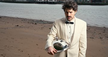 Paddy Young, a young white man with brown curly hair, a moustache, and stubbly beard dressed in a cream tuxedo on a beach with a town behind him. He is looking confused at the floor and holding a metal cloche
