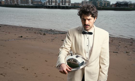Paddy Young, a young white man with brown curly hair, a moustache, and stubbly beard dressed in a cream tuxedo on a beach with a town behind him. He is looking confused at the floor and holding a metal cloche