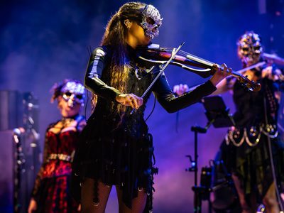 A woman in a sparkly skull mask and short black dress with skeletal sleeves is playing the violin with other musicians behind her also in matching skull masks