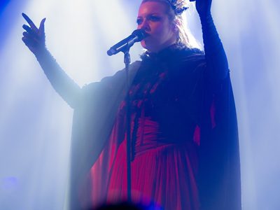 A woman in a long red dress and black cape with black lace gloves and tiara singing into a microphone with her arms raised