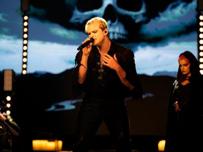 A man in a black tunic shirt with metal clips singing passionately into a microphone with a large skull on a screen behind him