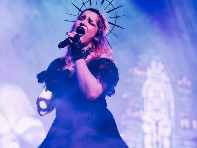 A woman in a large black lacey dress with fingerless lace gloves and a halo crown singing very passionately into a microphone