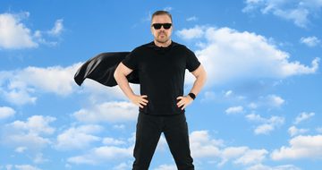 Ricky Gervais wearing a black T-shirt, jeans, sunglasses, and a cape, standing with hands on hips against a blue sky with clouds.