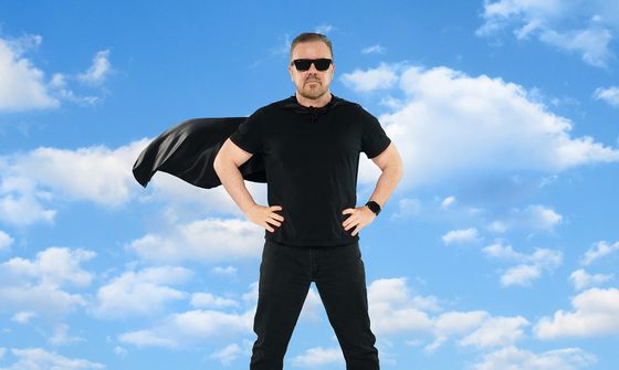 Ricky Gervais wearing a black T-shirt, jeans, sunglasses, and a cape, standing with hands on hips against a blue sky with clouds.