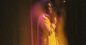 A hazy image of Richard Ayoade performing on stage in a yellow suit, and red tie holding a microphone