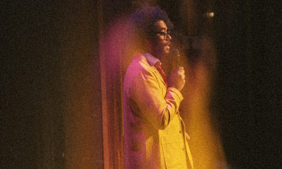 A hazy image of Richard Ayoade performing on stage in a yellow suit, and red tie holding a microphone