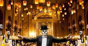 A man with his face painted like a skull in a suit and top hat holding a conductors baton with his arms wide spread in a grand hall surrounded by candles and skulls