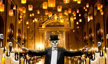 A man with his face painted like a skull in a suit and top hat holding a conductors baton with his arms wide spread in a grand hall surrounded by candles and skulls