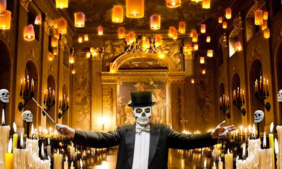 A man with his face painted like a skull in a suit and top hat holding a conductors baton with his arms wide spread in a grand hall surrounded by candles and skulls
