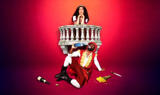 a girl with long curly dark hair leans on a balcony. Below her is a black man on the floor dribking from a gin bottle. Around him is a bunch of flowers, knife and a bottle of wine