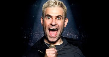 Simon Brodkin with his eyes and mouth very wide open staring down the camera under a spotlight holding a microphone